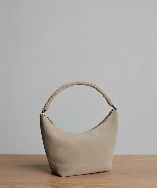 The Medium Crescent Bag in beige suede, made in Italy, features a curved shape and thick twisted handle. It is displayed on a light wooden surface with a plain gray background.
