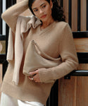 A woman in a cozy camel sweater with a lighter layer draped over her shoulders holds the Medium Crescent Bag. She stands indoors by a wooden wall, gazing to the side with a relaxed expression.