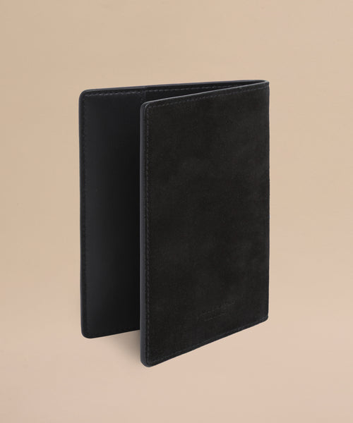 The Suede Passport Holder in black stands upright against a plain beige background, highlighting its soft texture and minimalist design—perfect for organizing your travel essentials.