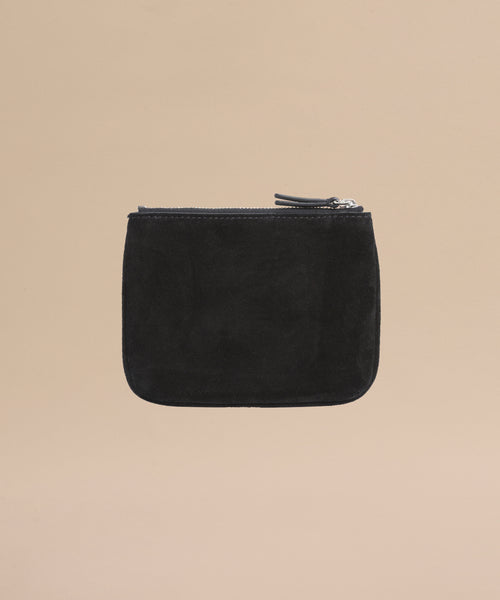 The Suede Pouch is a small black suede accessory with a zippered closure and minimalist design, perfect for use as a compact pouch or passport holder, pictured on a plain beige background.