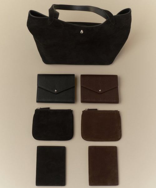 A black tote bag is showcased above six leather goods: two envelope wallets, two Suede Pouches in black and brown, and two slim rectangular card holders—all designed for stylish organization.