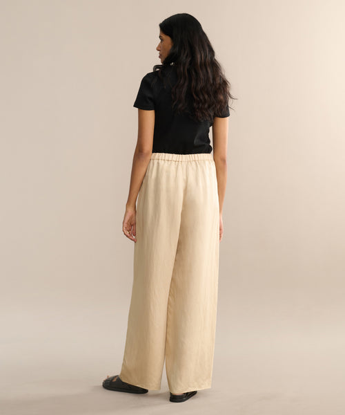 A woman with long, wavy dark hair stands facing away against a light background, wearing a black short-sleeve top, black sandals, and the Summer Demi Pant—loose linen pants.