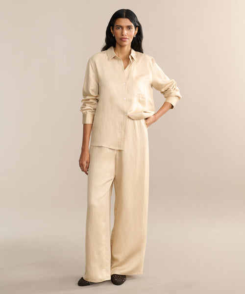 A woman stands against a plain beige background, wearing the Summer Demi Pant and a loose, light beige button-up shirt with sleeves pushed up. She has long dark hair and wears dark flat shoes.