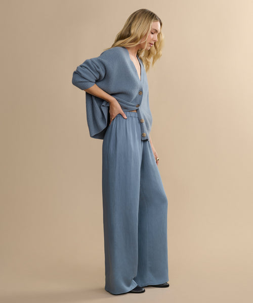 A woman with long blonde hair stands against a beige background, wearing a loose blue cardigan with large buttons, the Summer Demi Pant in linen texture, and black shoes. She looks down with a relaxed posture.