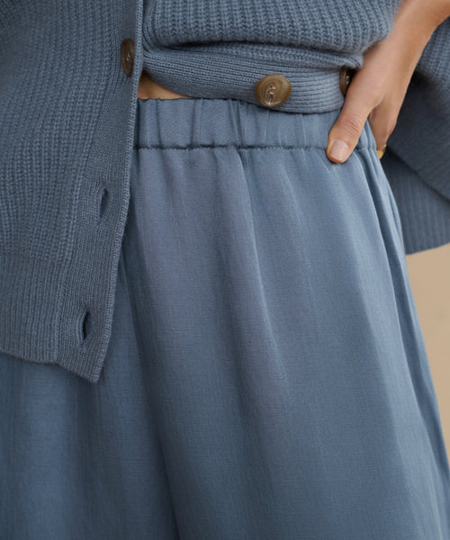 Close-up of a person wearing the Summer Demi Pant and a blue knit sweater with brown buttons, both showcasing a subtle linen texture, with one hand resting on their hip.