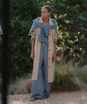 A person stands outdoors on a gravel path, wearing the Summer Demi Pant—a loose blue pant with a subtle linen texture—under a beige trench coat and a blue sweater draped over their shoulders. Green foliage and ornamental grasses fill the background.
