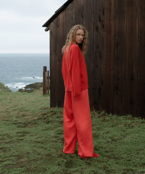 Woman wearing a bright red outfit standing in front of a wooden cabin by the ocean.