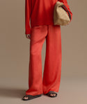 Woman wearing a red outfit with wide-leg pants and holding a woven bag against a beige background