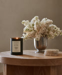 A hand-poured Sun Valley Glass Candle in black sits on a wooden table next to a metal vase with white flowers and round coasters, with a softly blurred background.