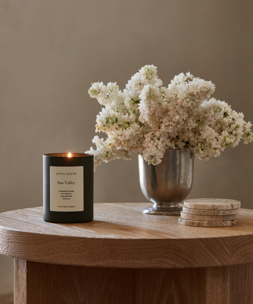 A hand-poured Sun Valley Glass Candle in black sits on a wooden table next to a metal vase with white flowers and round coasters, with a softly blurred background.
