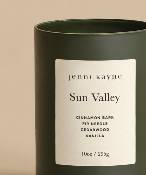 The Sun Valley Glass Candle is a 10oz hand-poured dark green glass candle with a beige label reading jenni kayne Sun Valley. Notes: Cinnamon Bark, Fir Needle, Cedarwood, and Vanilla. Shown on a light beige background.