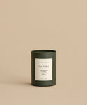 A dark green, hand-poured Sun Valley Glass Candle sits on a beige surface. Its white label highlights notes of Cinnamon Bark, Cedarwood, Vanilla, and states 10oz / 283g against a neutral background.
