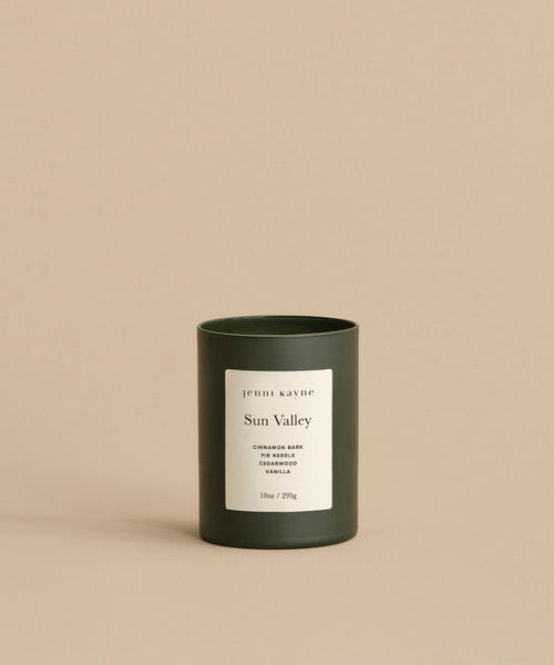 A dark green, hand-poured Sun Valley Glass Candle sits on a beige surface. Its white label highlights notes of Cinnamon Bark, Cedarwood, Vanilla, and states 10oz / 283g against a neutral background.