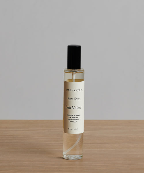 A clear Sun Valley Room Spray bottle with a black cap rests on wood against a plain gray background. The label highlights scents of cinnamon bark, cedarwood, vanilla, and balsam fir for a warm seasonal refresh.