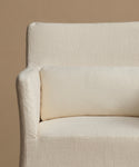 Close-up of a cream-colored upholstered armchair with a matching lumbar pillow, set against a solid warm beige background.