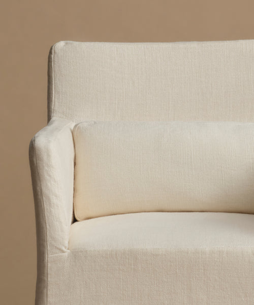Close-up of a cream-colored upholstered armchair with a matching lumbar pillow, set against a solid warm beige background.