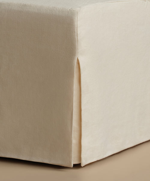 Close-up of a beige upholstered ottoman or bench corner with a skirted fabric cover and a front slipper opening, shown against a matching neutral background.