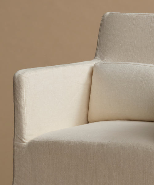 Close-up of a cream-colored upholstered armchair with a textured linen-like fabric, featuring a square armrest and a matching back cushion against a solid warm beige background.