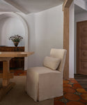 A beige upholstered dining chair with a matching back cushion in a rustic, warm-toned room with terracotta tile flooring and a wooden table.