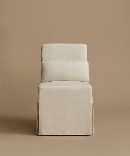 An upholstered accent chair with a tiered backrest and a skirted base, covered in a textured cream fabric, set against a solid warm beige background.