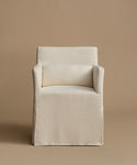 A slipcovered armchair in a warm off-white linen fabric, featuring a matching rectangular lumbar pillow, set against a solid beige studio background.