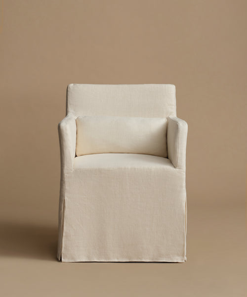 A slipcovered armchair in a warm off-white linen fabric, featuring a matching rectangular lumbar pillow, set against a solid beige studio background.