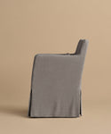 A side view of the Sunset Dining Arm Chair, featuring a gray upholstered slipcover with a long skirt concealing the legs, set against a plain beige background.