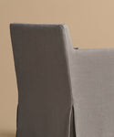 A close-up view of the back and side of the Sunset Dining Arm Chair, featuring modern gray linen upholstery and a tailored skirt, set against a plain beige background.
