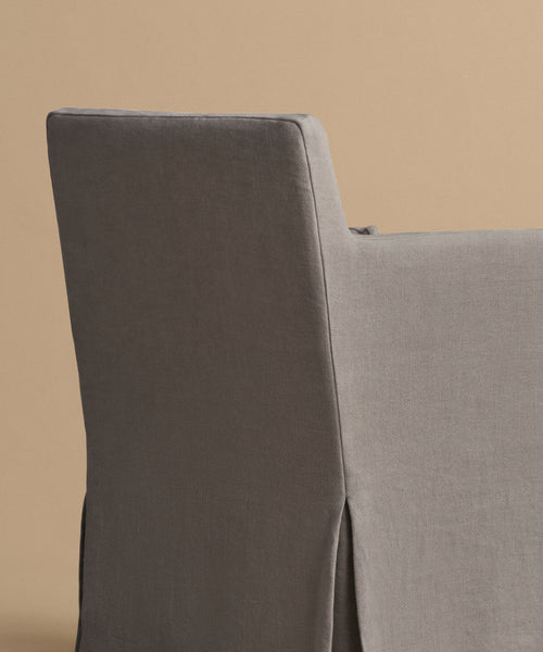 A close-up view of the back and side of the Sunset Dining Arm Chair, featuring modern gray linen upholstery and a tailored skirt, set against a plain beige background.