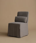 A single upholstered accent chair in a dark gray fabric slipcover, featuring a high back, a matching rectangular back cushion, and a skirted base, set against a solid warm beige studio background.