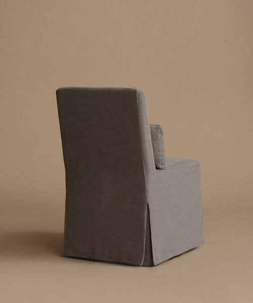 A single upholstered armchair shown from the back in a studio setting, with a tall back, slim arms, and a skirted base. The chair is covered in a textured fabric in a warm gray-brown tone against a solid beige backdrop.