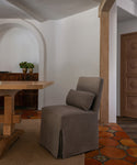 A gray upholstered dining chair with a matching cushion in a dining room with terracotta tile flooring, a wooden table, and white walls.