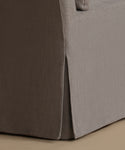 Close-up of a slipcovered upholstered furniture corner, showing a taupe-brown side panel and a light gray front skirt with a slight drop shadow on a beige floor.
