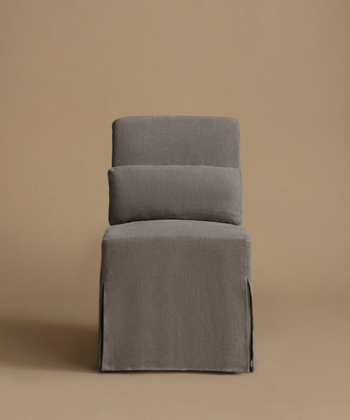 A gray upholstered chair with a high back, two loose back cushions, and a skirted base, photographed against a solid warm beige studio background.