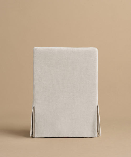 The Sunset Dining Arm Chair is shown from behind, featuring a beige linen tailored slipcover with neat seams and side vents, and a straight back, set against a light tan background.