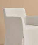 The Sunset Dining Arm Chair is upholstered in light beige with a tailored slipcover, soft fabric, straight arms, and a gently curved backrest. It offers a modern, minimal look and is shown against a plain tan background.