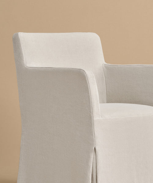 The Sunset Dining Arm Chair is upholstered in light beige with a tailored slipcover, soft fabric, straight arms, and a gently curved backrest. It offers a modern, minimal look and is shown against a plain tan background.