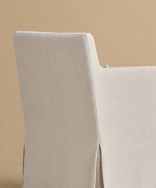 The Sunset Dining Arm Chair, featuring a beige linen slipcover and straight backrest, is set against a plain light brown background.