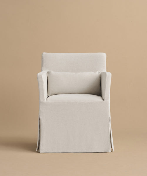 The Sunset Dining Arm Chair, shown front-facing against a plain beige background, features a high back, light beige linen upholstery, straight armrests, a matching lumbar pillow, and a skirted bottom for an elegant look.