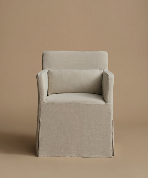 A slipcovered armchair with a matching back cushion, upholstered in a light beige textured fabric, set against a solid warm beige background.