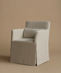 A slipcovered armchair with a matching back cushion in a light oatmeal linen fabric, set against a solid warm beige studio background.