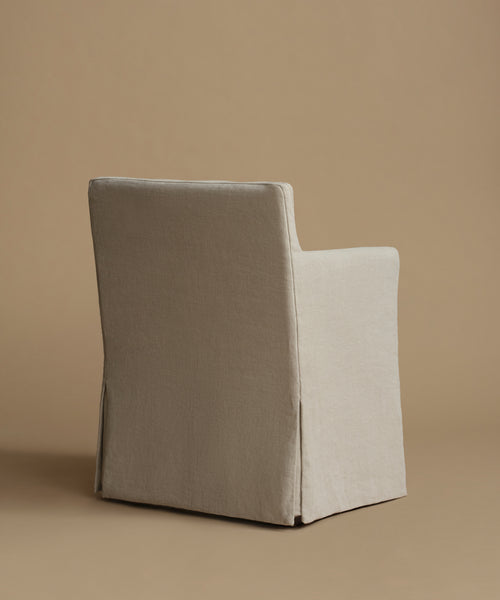 A single upholstered armchair shown from the back, covered in a light warm gray-beige fabric, set against a solid tan studio background.