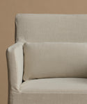 Close-up of a light beige upholstered armchair with a matching rectangular lumbar pillow against a solid warm beige background.