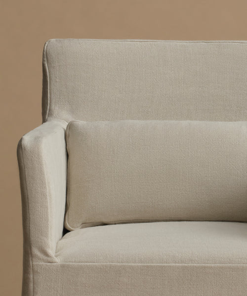 Close-up of a light beige upholstered armchair with a matching rectangular lumbar pillow against a solid warm beige background.