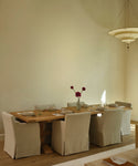 A rustic wooden dining table set with linen-covered chairs in a warm, minimalist dining room with beige walls and a hanging pendant light.