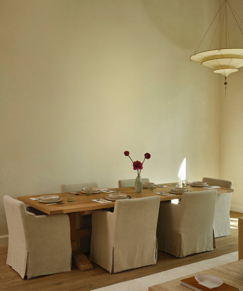 A rustic wooden dining table set with linen-covered chairs in a warm, minimalist dining room with beige walls and a hanging pendant light.