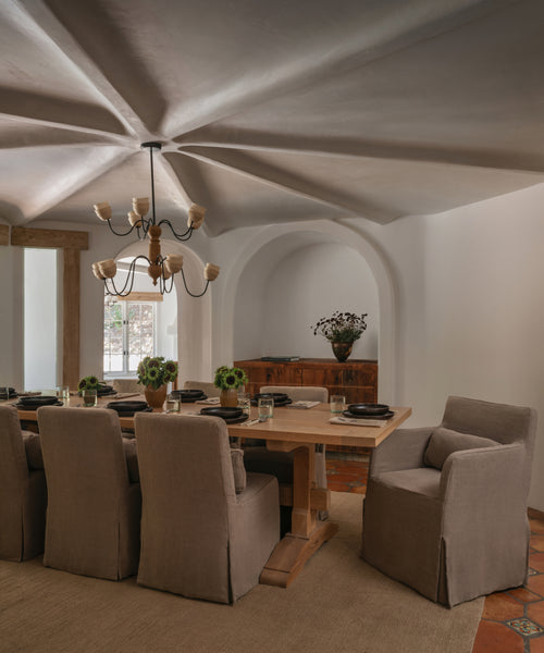 A styled dining room interior featuring a wooden dining table surrounded by upholstered dining chairs in a neutral beige fabric, set under a decorative starburst plaster ceiling and an arched alcove background.