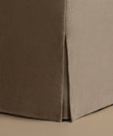 Close-up of a slipcovered upholstered corner of a piece of furniture, showing a taupe-brown side panel and a light taupe front panel with a vertical seam, set against a matching beige background.
