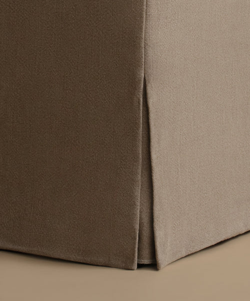 Close-up of a slipcovered upholstered corner of a piece of furniture, showing a taupe-brown side panel and a light taupe front panel with a vertical seam, set against a matching beige background.
