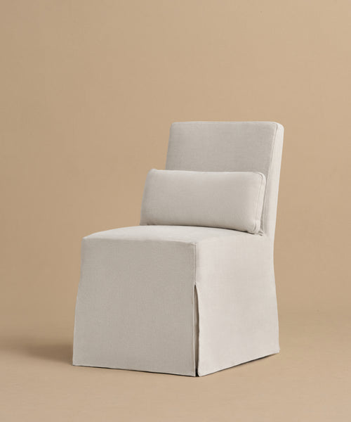 The Sunset Dining Chair, with light beige upholstery and a matching rectangular lumbar pillow, features a modern, boxy California-inspired design and draped fabric cover. It is shown here on a plain beige background.
